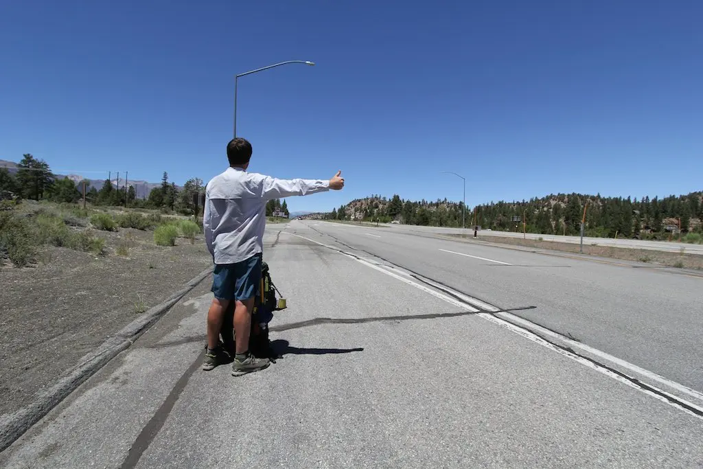 A person with short hair, wearing a long-sleeve shirt, shorts, and boots, stands by an empty highway under a clear blue sky. Theyre hitchhiking with their thumb up and a backpack on the ground beside them. Sparse greenery lines the road—a scene reminiscent of the Pacific Crest Trail.