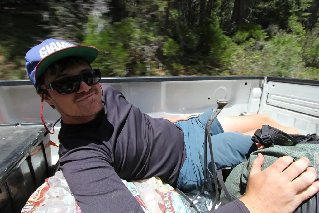 A man in sunglasses and a cap lies relaxed in the back of a moving pickup truck, embodying the spirit of a long hitch. Clad in shorts and a long-sleeve dark shirt, he clutches his backpack as the lush greenery blurs by, reminiscent of scenic hikes through Yosemite.