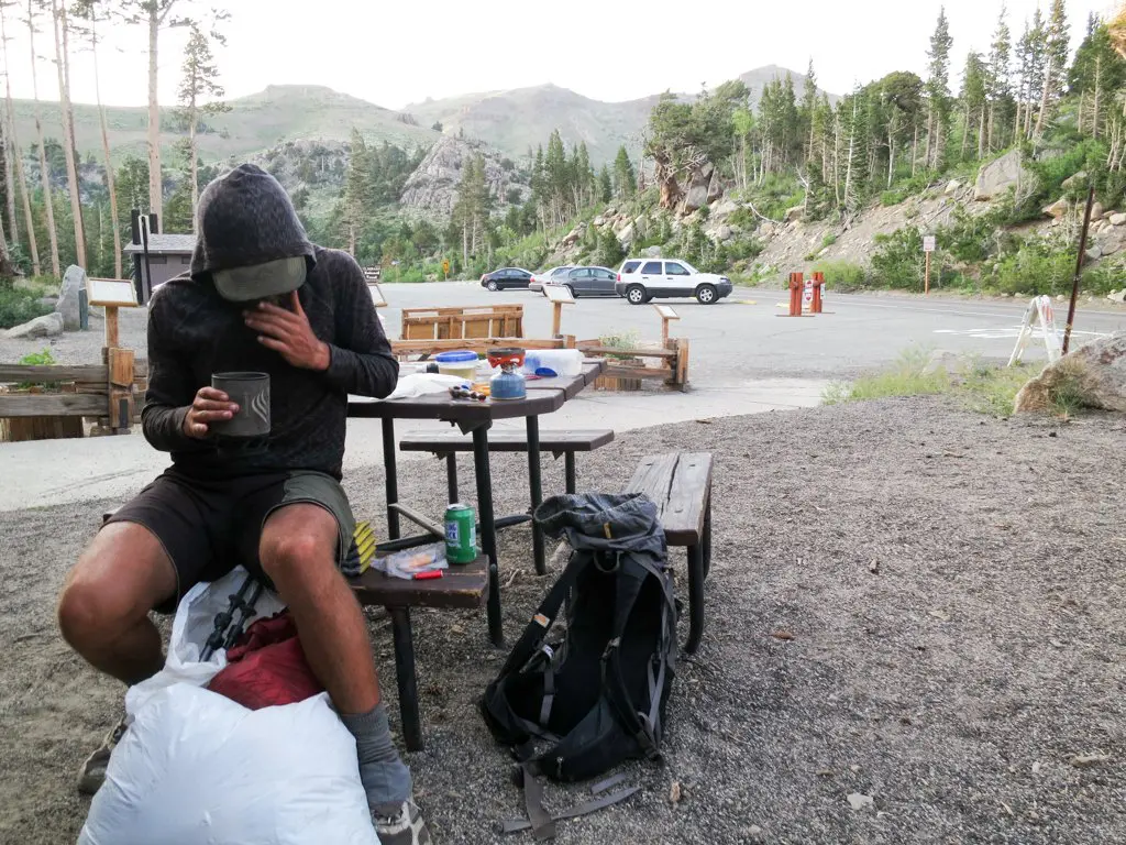 A person in a hoodie sits at a picnic table, savoring a meal in the mountainous, wooded area of the Pacific Crest Trail. A backpack and white bag are on the ground beside them. Nearby, two cars are parked under the canopy of trees and hills. Its a casual outdoor campsite setting.