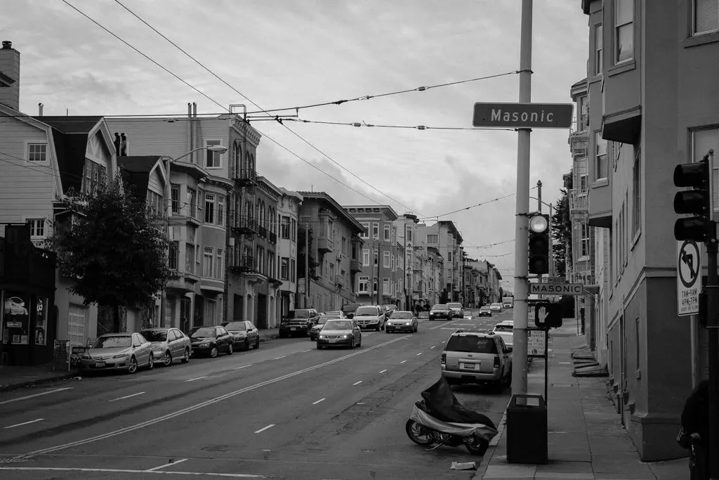 A black and white artsy photo captures a street in an urban area, lined with parked cars. A few pedestrians stroll along the sidewalk beneath overhead wires for streetcars. A street sign reading Masonic adds a touch of intrigue to this artful scene, perfect for any photo gallery.