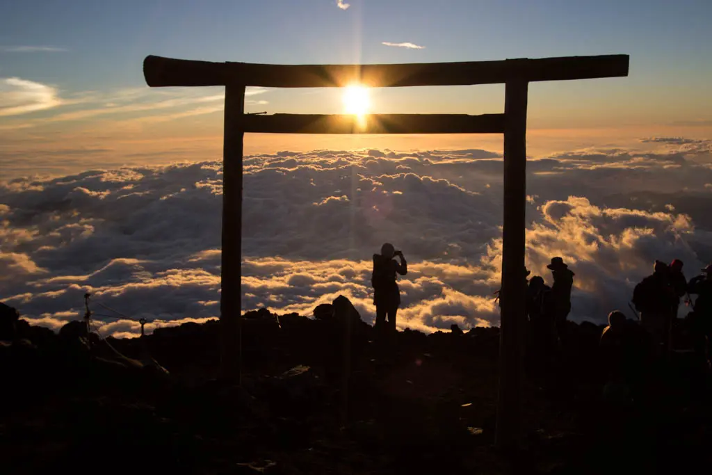 Silhouetted people stand near a torii gate atop a mountain, with the sun setting behind the horizon. Below, a sea of clouds creates a dramatic scene over Mount Fuji, blending warm and cool colors in the sky.