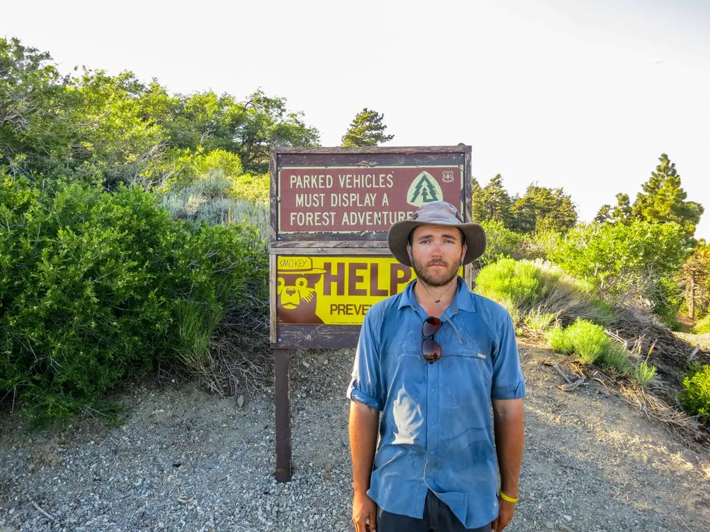 A person wearing a wide-brimmed hat and blue shirt stands in front of a trailhead sign at a forest location. The backdrop features greenery and a partly cloudy sky. The sign, which might spark questions, mentions parking permits and the Pacific Crest Trail name.