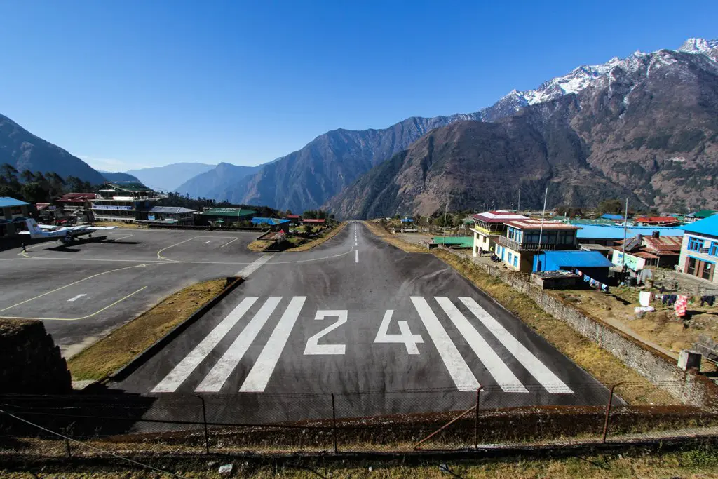 A sloped runway with the number 24 painted on it at a mountain airport near Everest Base Camp. Surrounding buildings boast colorful roofs, while snow-capped peaks and a clear blue sky provide a breathtaking backdrop.