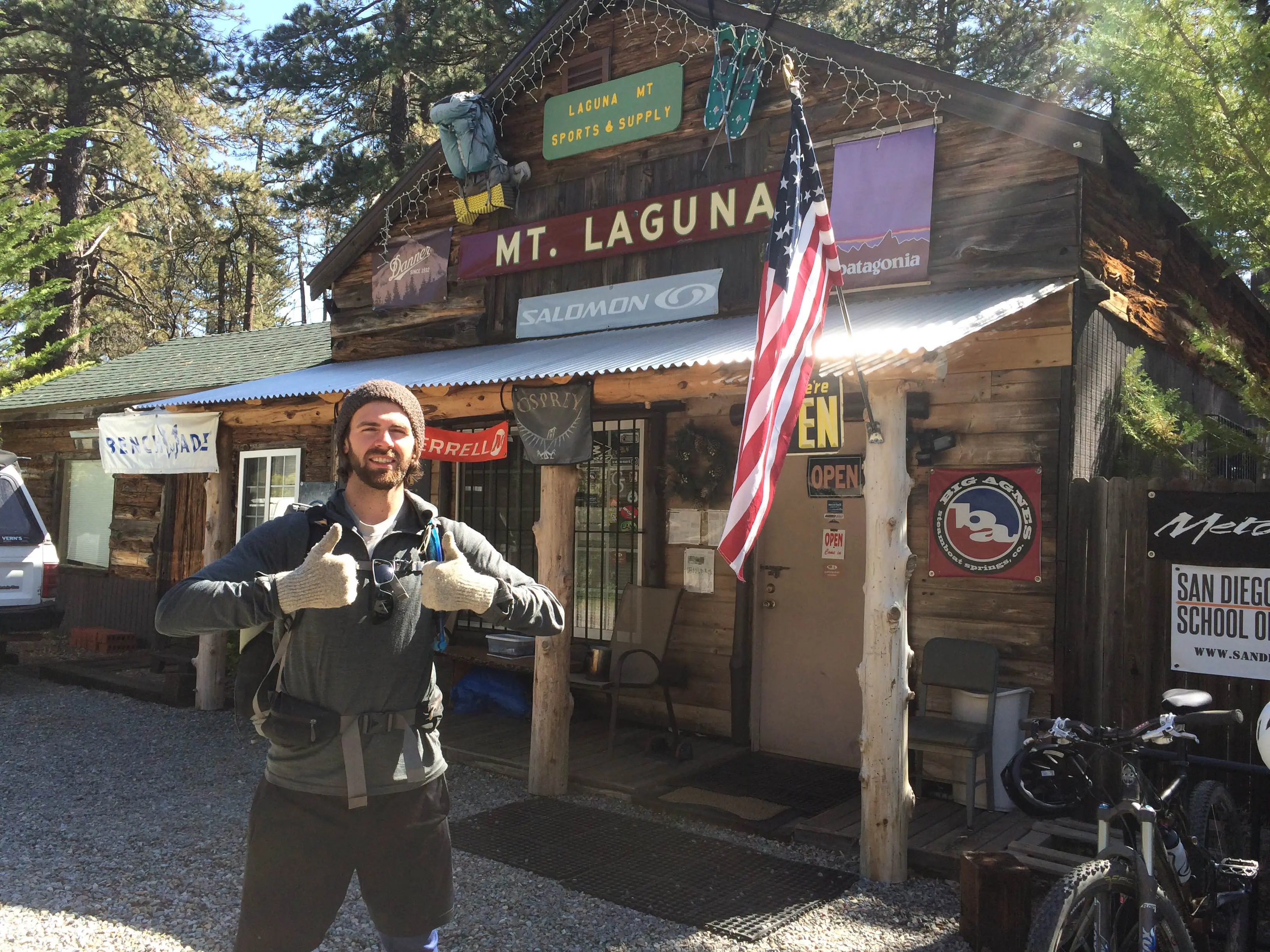 A person stands smiling in front of a rustic wooden shop labeled Mt. Laguna, reminiscent of the classic Mount Laguna Outfitter. Theyre giving a thumbs-up, with an American flag waving and bicycles parked out front. Its a sunny day, embraced by natures leafy embrace.