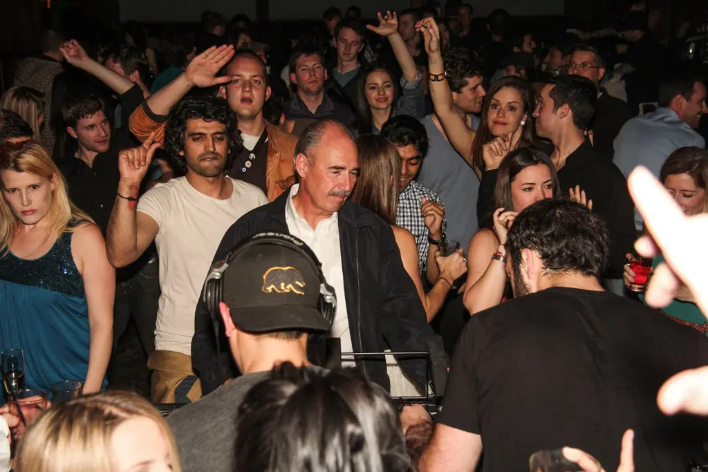 A lively crowd dances at a party, with a DJ in the foreground wearing a cap. People of diverse ages are enjoying the music, with hands in the air and smiles all around. The atmosphere is vibrant and social, embodying a spirit of diversity and unity as contributors to an unforgettable night.