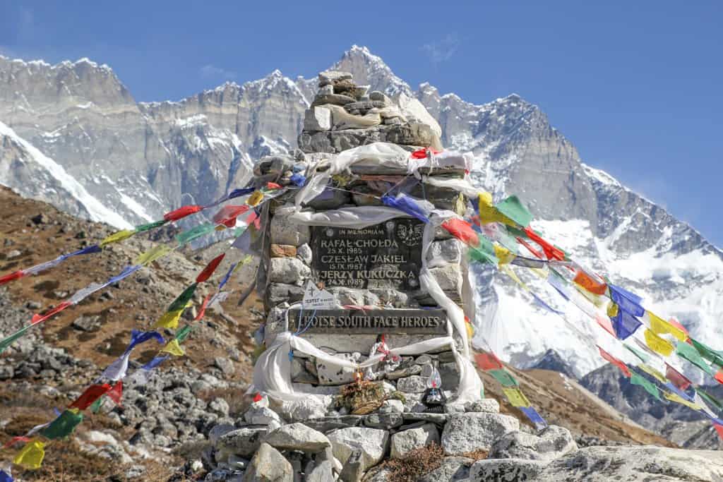 A stone memorial adorned with colorful prayer flags stands against a backdrop of snowy mountains near Everest Base Camp. The plaque honors individuals who contributed to a historic event, under a clear blue sky overhead.
