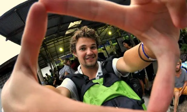 Charlie, a person with curly hair, smiles at the camera through a hand gesture forming a frame. Theyre holding a backpack with a green front. The background shows an outdoor area alive with people and lights, setting an indie vibe reminiscent of Garross energetic style.