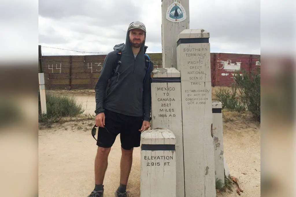 A person stands by the monument marking the southern terminus of the Pacific Crest National Scenic Trail, jotting notes in their trail journal. The sign details trail distance, elevation, and establishment date. Behind them, a fence stretches across the vast desert landscape under a clear sky.