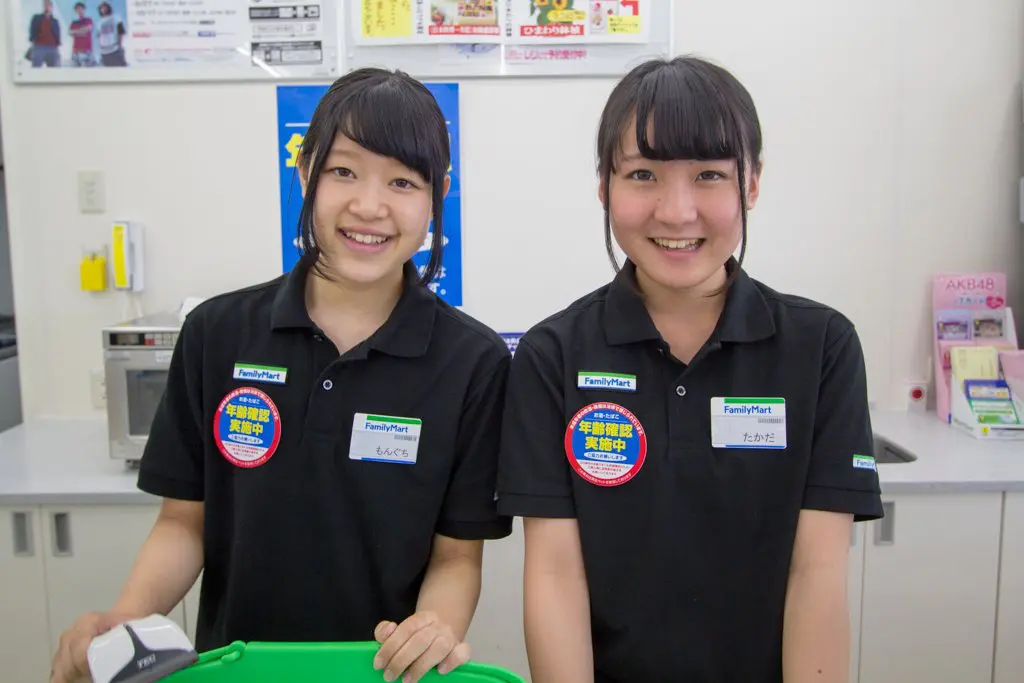 Two young women in black uniforms stand smiling behind the konbini counter. They are working at FamilyMart, as indicated by their name tags. A green shopping basket is partially visible in front of them, with store signage and shelves filling the background.