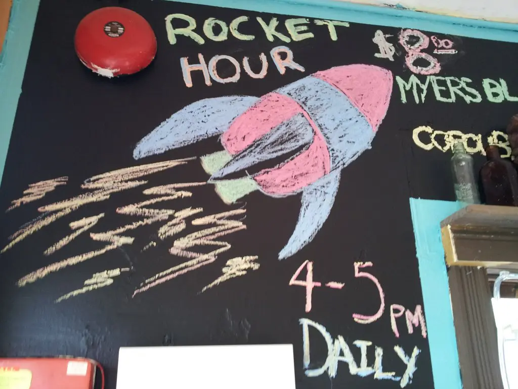 A chalkboard drawing features a colorful rocket blasting off with Rocket Hour text, hinting at a secret rocket fuel recipe. It advertises 4-5 PM Daily for $8, mentions Myers Bl, and includes the partial word, Co.... Bright chalk hues like pink, blue, and yellow enhance the charm.