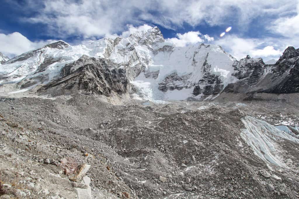 The snow-covered mountainous landscape resembles Everest Base Camp, with its rugged terrain and rocky slopes under a partly cloudy sky. The sharp, steep peaks reveal patches of ice and snow, conveying a sense of vast, untouched wilderness.