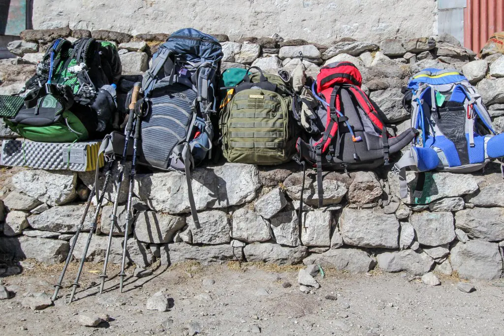 Five colorful hiking backpacks and trekking poles rest against a sunlit stone wall in Duglha. The backdrop is rustic, and the ground is dusty. These packed bags boast a variety of vibrant colors and designs, ready for an adventurous journey.