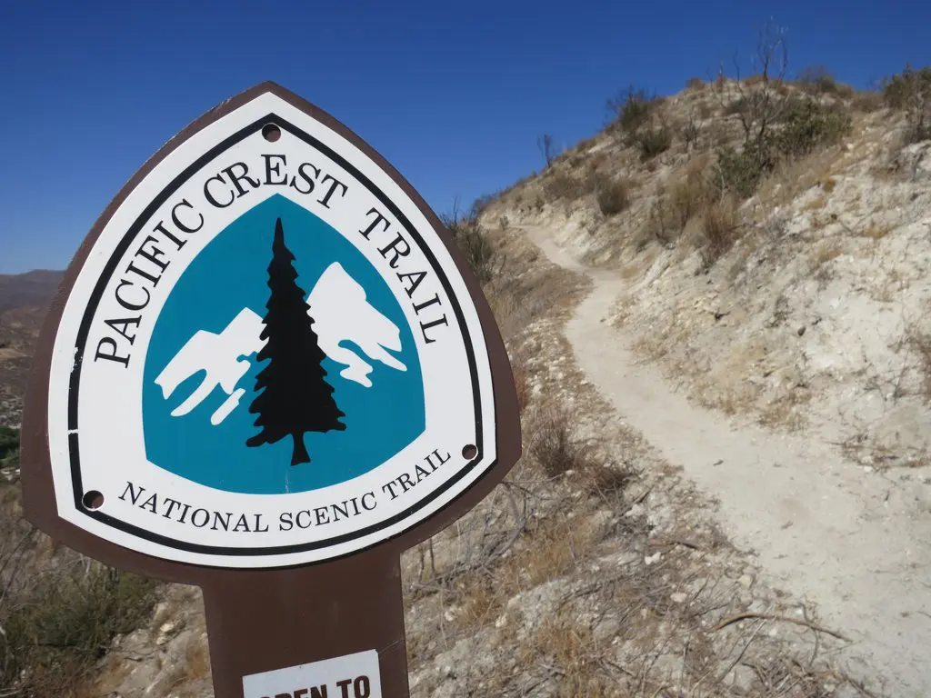 A close-up of the Pacific Crest Trail sign with a tree and mountain logo marks the start of a narrow dirt path. The trail winds through dry, rocky terrain under a clear blue sky, inviting adventurers to explore its breathtaking vistas.