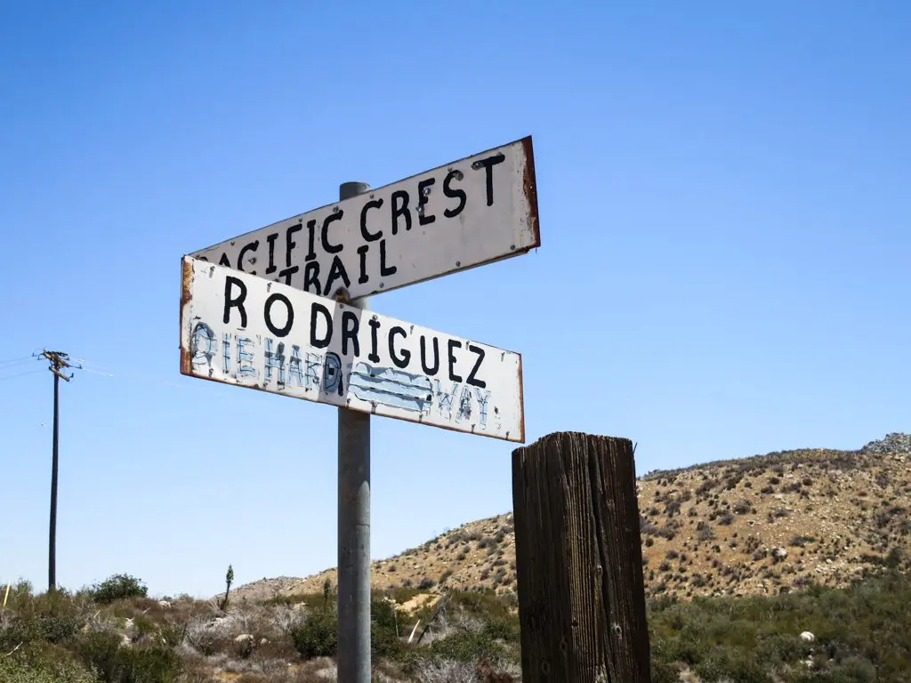 Weathered directional signs stand against a clear blue sky. The top sign, proudly pointing left, reads Pacific Crest Trail, while the bottom sign, partly obscured but visible, directs right to Rodriguez, all framed by the vast desert landscape in the background.
