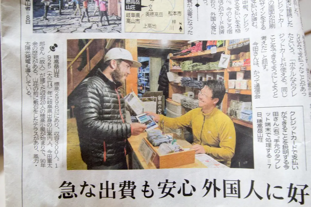 A person in a black jacket and cap stands at a shop counter, engaging with a smiling shopkeeper in a yellow shirt. Shelves brimming with products frame the scene, Japanese text weaving through the image like trails in the Kita Alps.