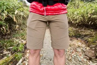 A person in a red jacket and khaki shorts stands on a rocky path, their lower body framed by verdant greenery and a chockstone adding rugged charm to the scenic backdrop.