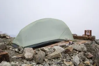 A gray tent, branded with SuperMegaUL 2, is pitched on a rocky mountain ledge under a cloudy sky. Large stones and wooden beams secure the tent firmly. A sign displaying the number 23 is visible on the right side.