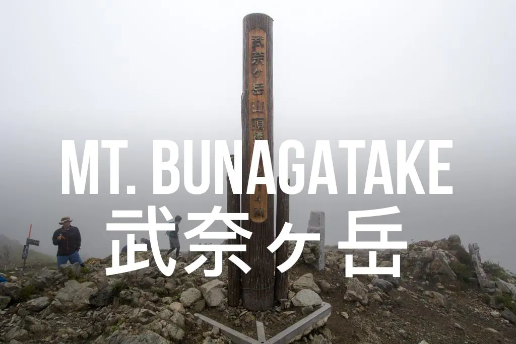 Foggy mountain summit with a wooden post displaying Mt. Bunagatake in English and Japanese. Nearby, two people pause at the rocky terrain, their thoughts perhaps wandering to distant peaks like Mount Tate.