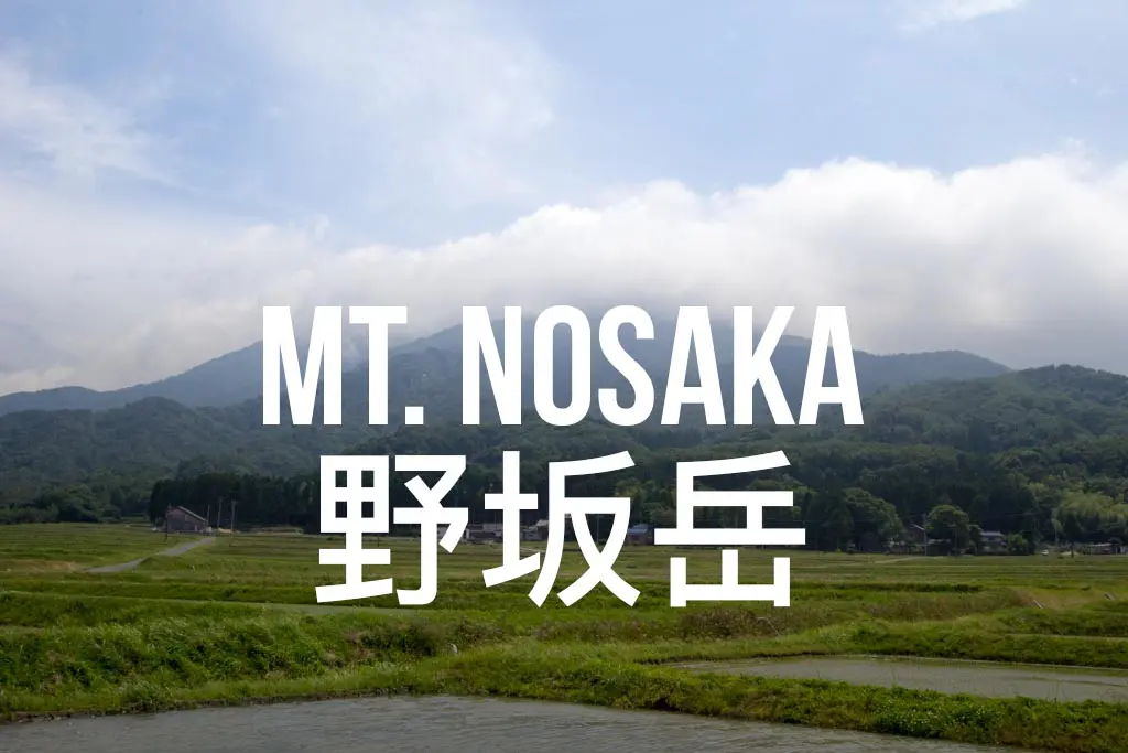 In this captivating image, Mt. Nosaka graces the backdrop, while lush green fields stretch out beneath a partly cloudy sky. The scene invites comparisons to Mount Tate. Bold white text overlays the image, showcasing MT. NOSAKA alongside Japanese characters below.