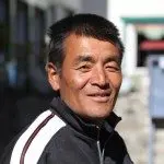 A person with short dark hair is smiling, wearing a black jacket with white and red stripes on the collar and sleeves. The slightly blurred background hints at the breathtaking scenery of Nepal, contrasting beautifully with their joyful expression.
