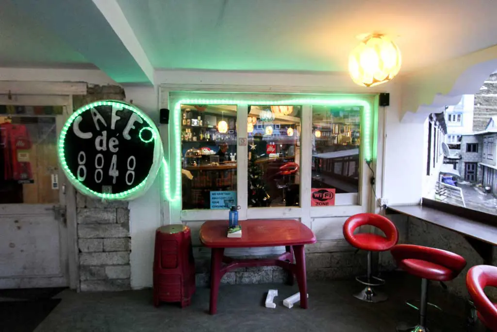 A cozy cafe interior with a green neon sign reading Cafe de 8848 reminiscent of Namche Bazaar. Theres a red table, red bar stools, and a window offering a view of the street outside. A Wi-Fi sign is visible by the window.