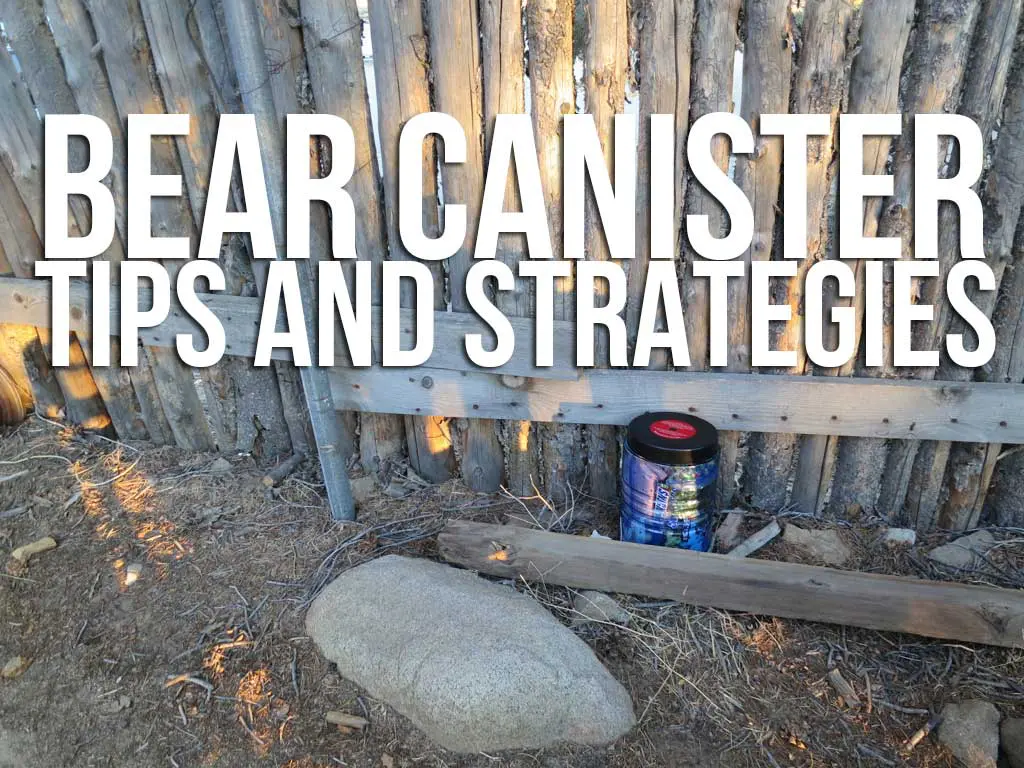A bear canister rests on the ground beside a rustic wooden fence, with large white text above proclaiming, Bear Canister Tips and Strategies. A rock and wooden plank complete the scene, inviting you to explore effective strategies for your outdoor adventures.