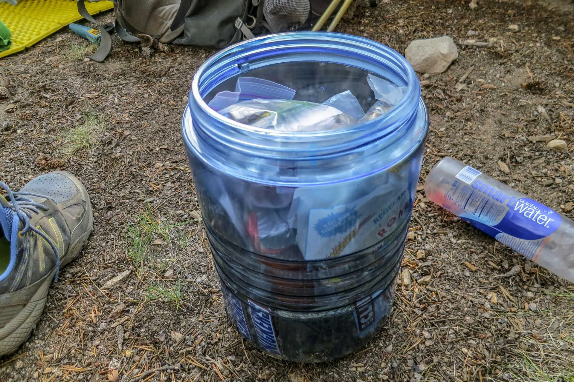A blue plastic bear canister, hailed as the best bear canister by seasoned hikers, sits among camping gear, filled with assorted food packages. Nearby, a clear water bottle glistens next to a rugged pair of hiking shoes.