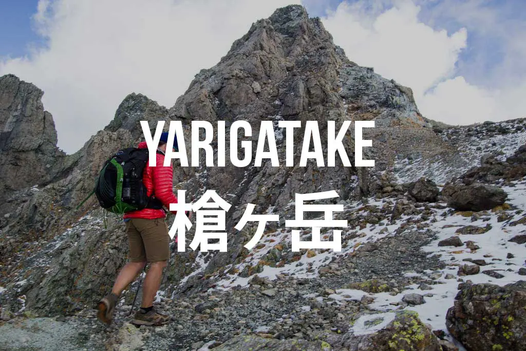 A person in a red jacket and shorts is hiking towards the rugged Yarigatake peak, its name elegantly overlaid alongside Japanese characters. Patches of snow dot the landscape beneath a cloudy sky.