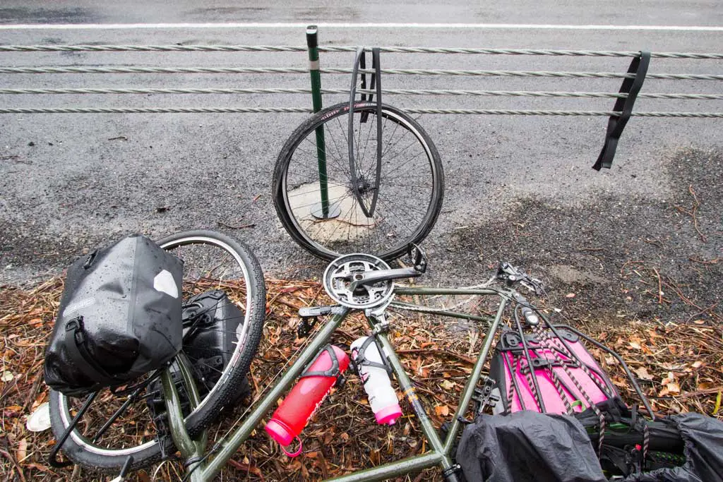 A disassembled bicycle lies on the ground beside a road with metal railings, perhaps from an unexpected flat tire during a bike tour day. The frame is in the foreground, wheels detached and leaning against the railing. Two water bottles cling to the frame, and a bag rests nearby.