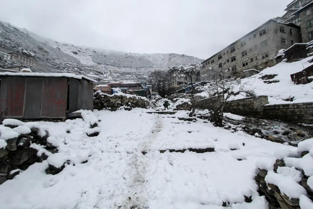 A snow-covered path leads through a mountain village, flanked by stone buildings and bare trees, reminiscent of Everest Base Camp on day 13. The mountainous backdrop is veiled in mist, enhancing the wintry scene.