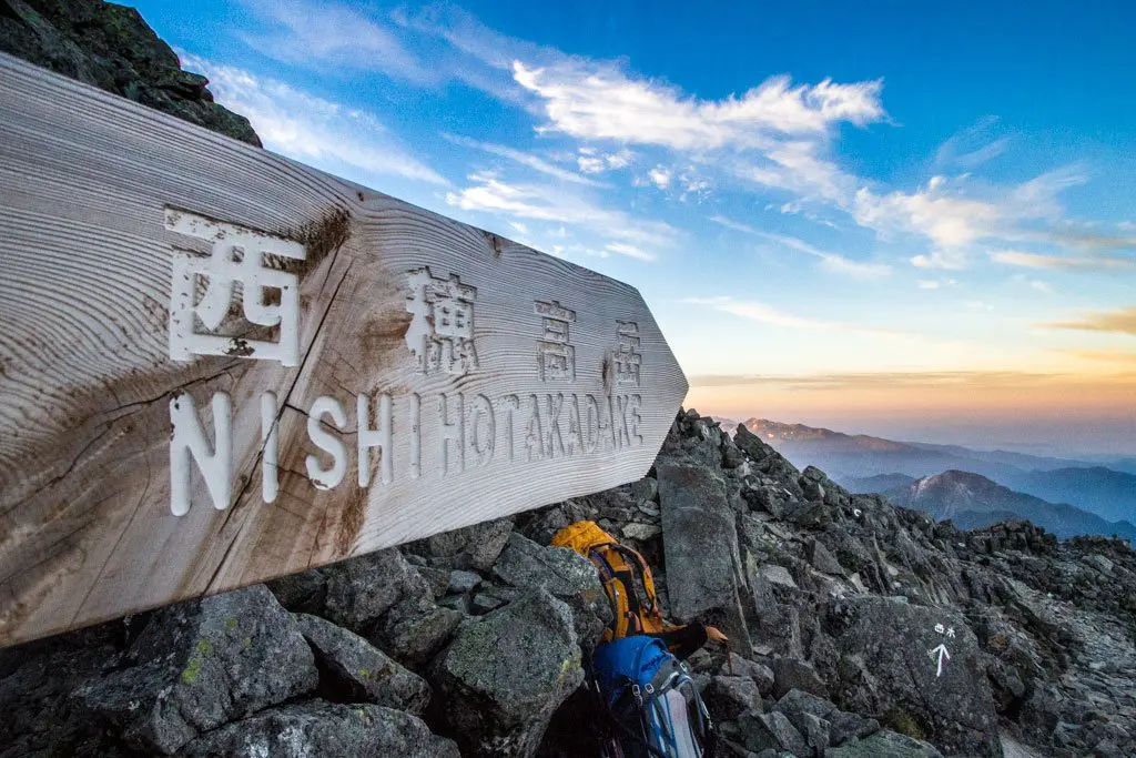 A wooden trail sign with Japanese and English text NISHI HOTAKADAKE stands in the rocky mountain landscape leading from Oku-Hotakadake. Nearby, a backpack rests as wispy clouds stretch across the vast sky.