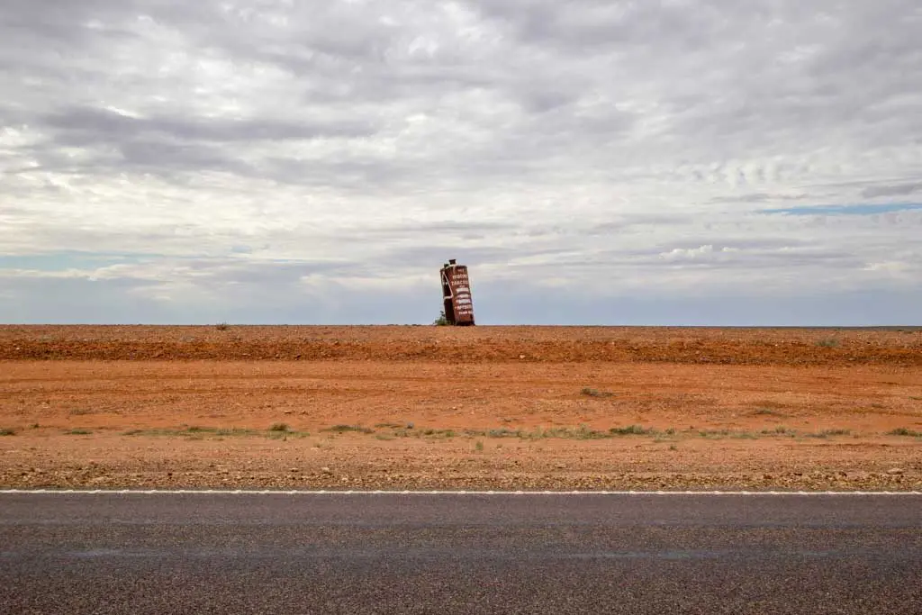 A rusted car is partially buried nose-first in the middle of a barren, flat desert landscape under a cloudy sky. The scene, discovered on Day 30 of the Australia Bike Tour, is framed by an empty road in the foreground.