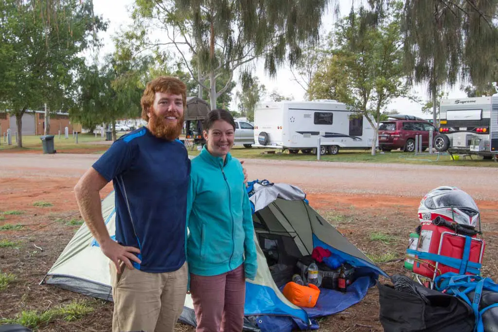 A man and woman smile, standing beside a tent at their campsite, as part of their Australia Bike Tour. Behind them are trees, parked cars, and camper trailers. Camping gear is scattered near the tent, both dressed in casual outdoor clothing. Its Day 34 of their adventure.