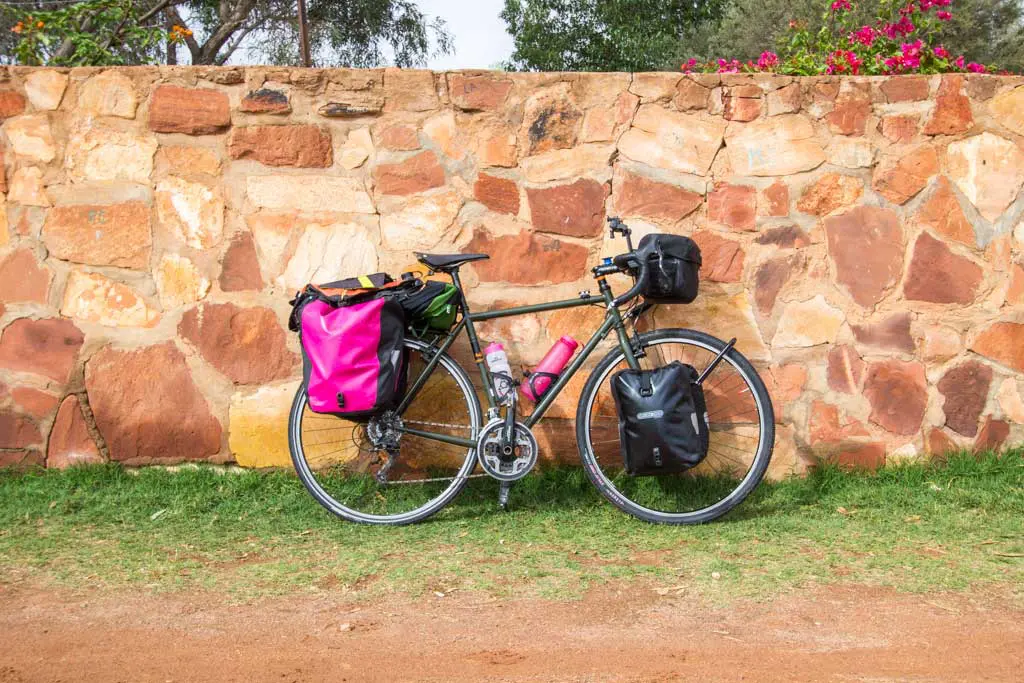 A touring bicycle with colorful panniers is parked against a rustic stone wall, reminiscent of an Australia Bike Tour. The bike has a pink water bottle on the frame and rests on grassy and dirt ground. Bright flowers bloom atop the wall in the background.