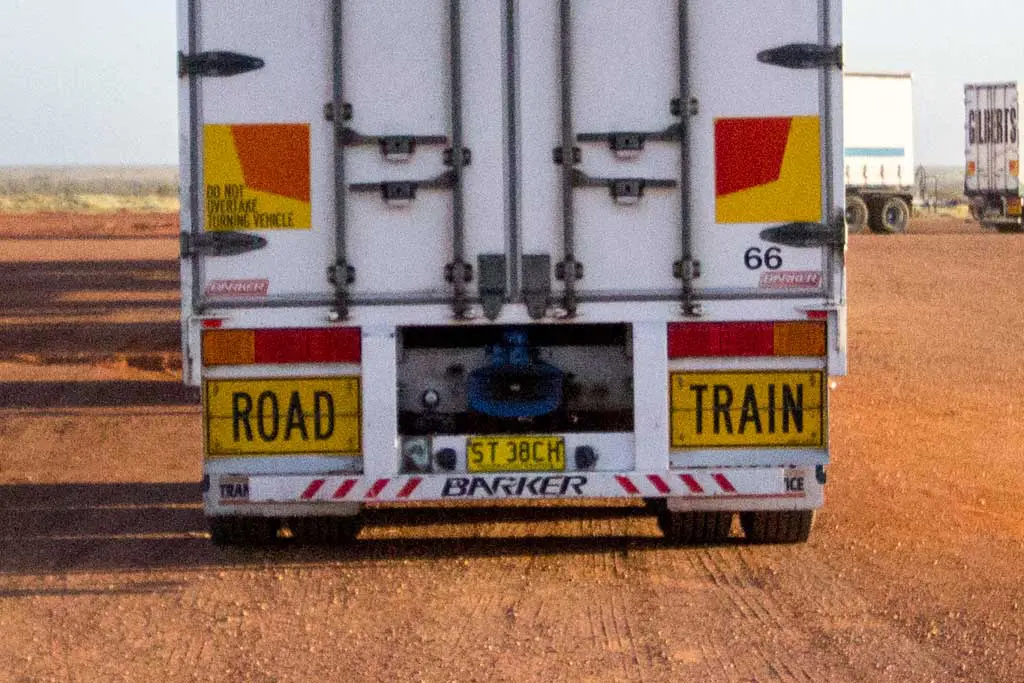 Rear view of a large ROAD TRAIN truck on a dusty Australian road, perfect for an adventurous Australia bike tour. The trucks dual rear doors feature safety stripes and warning signs, with a blue hitch underneath and another vehicle visible in the distance.