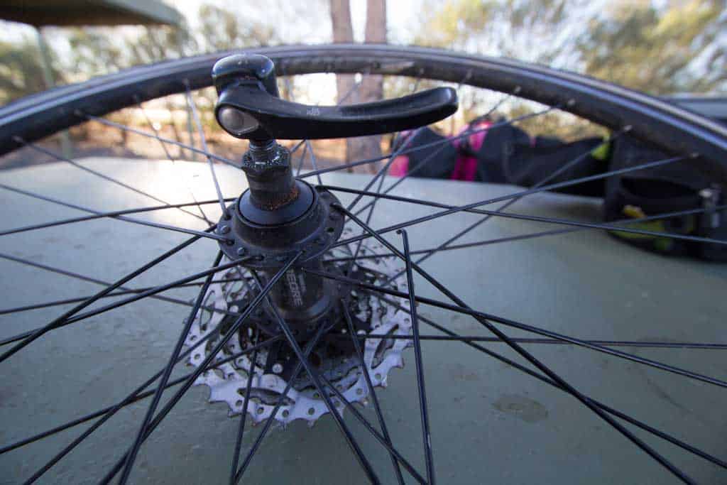 Close-up of a bicycle wheel lying on its side, showcasing the hub and gear cassette. A blurred backdrop hints at trees from the Australia Bike Tour on Day 50, capturing a moment where even a broken spoke couldnt dampen the adventure.