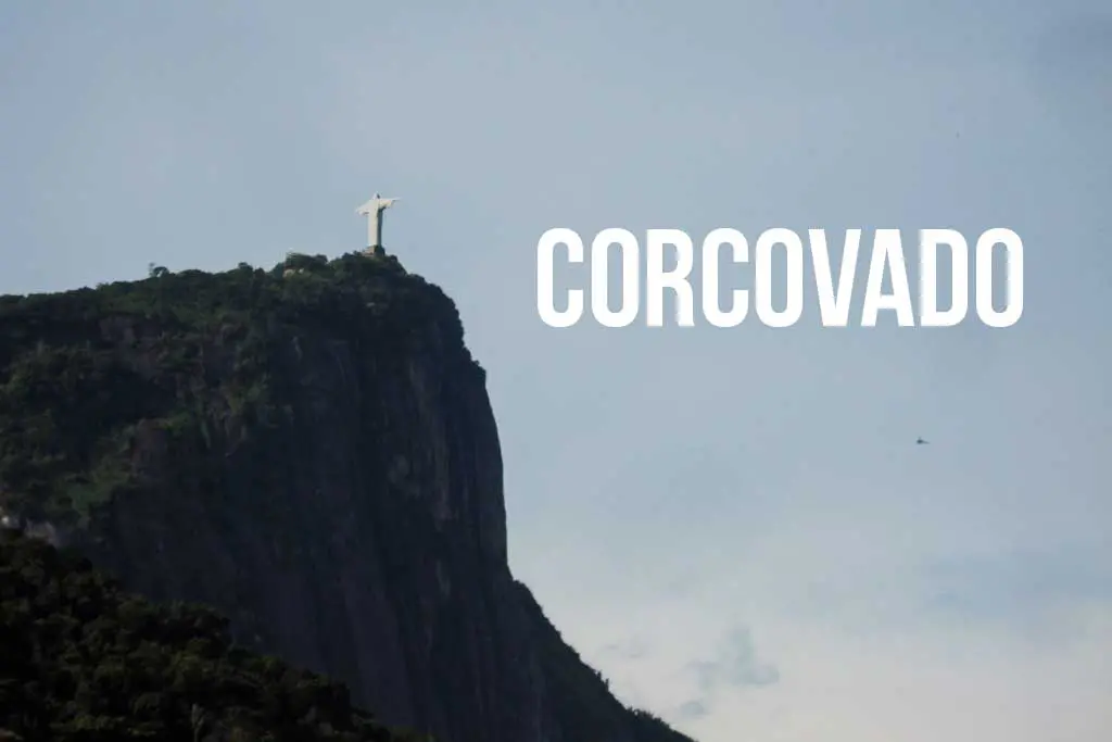 The image showcases the iconic Christ the Redeemer statue gracefully perched atop Corcovado Mountain in Rio de Janeiro, Brazil, under a clear sky. The word CORCOVADO is elegantly overlayed on the sky to the right of the statue.