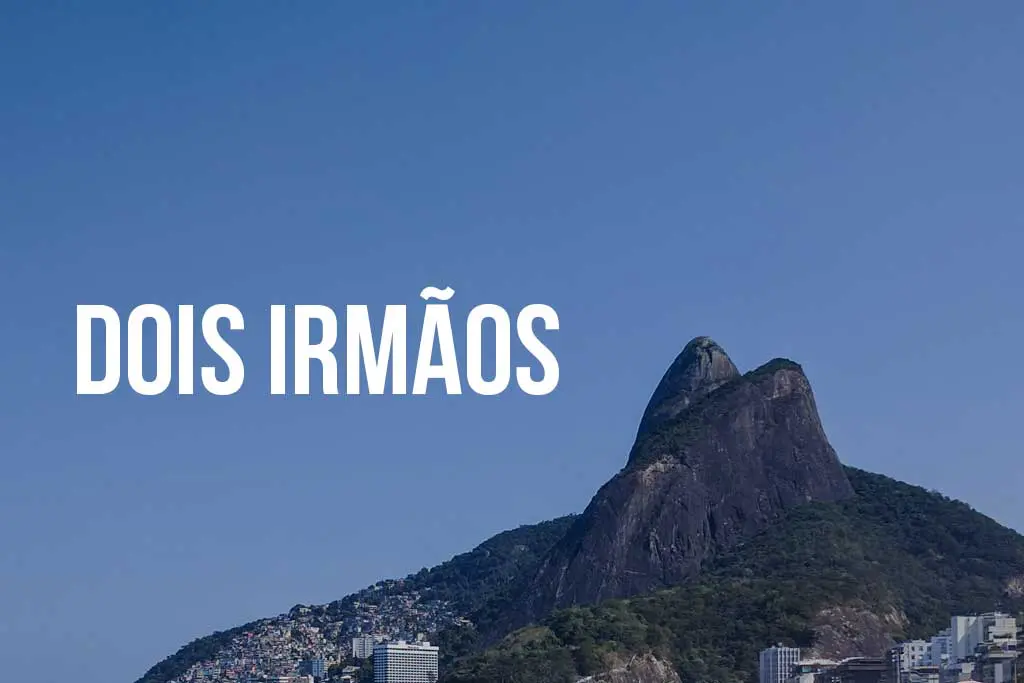 A clear blue sky frames the majestic Dois Irmãos mountain peak. Lush green foliage and a charming town nestle at its base. Bold white text reading DOIS IRMÃOS overlays the picturesque scene, highlighting the natural beauty.