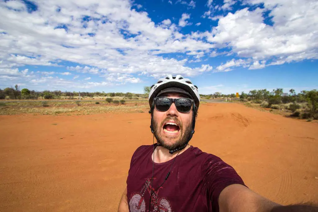 A person with a wide, open-mouthed smile takes a selfie in a vast, arid landscape wearing sunglasses and a helmet. A tailwind seems to lift their spirits as they sport a maroon shirt under the partly cloudy sky, looking cheerful and ready for adventure.