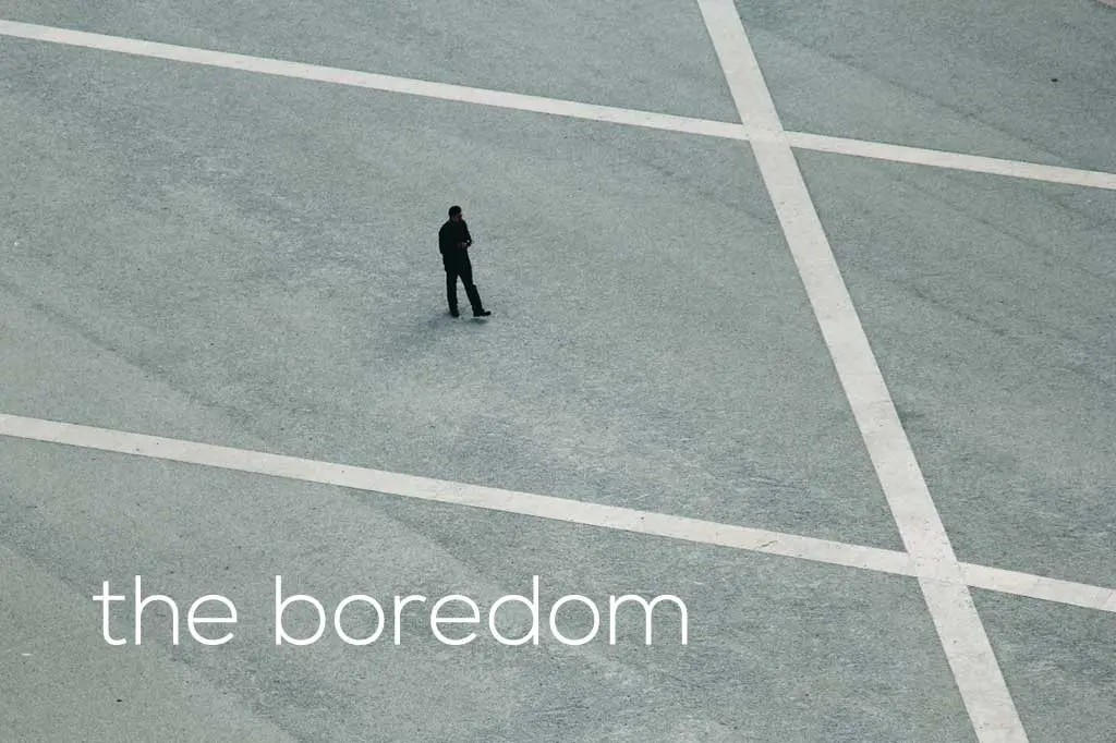 A solitary figure stands on a large, empty concrete surface intersected by white lines. The person appears small against the vast expanse. Text overlay reads, the boredom is palpable on Day 58 of the Australia Bike Tour.