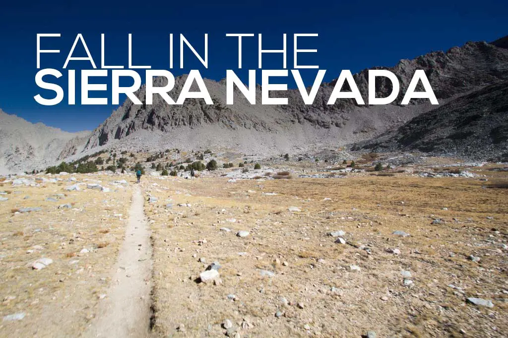 A dry Sierra Nevada landscape unfolds under a clear blue sky, with a narrow dirt trail winding through the rugged terrain. Sparse vegetation peeks out among rocks and grass patches. FALL IN THE SIERRA NEVADA is elegantly inscribed at the top.
