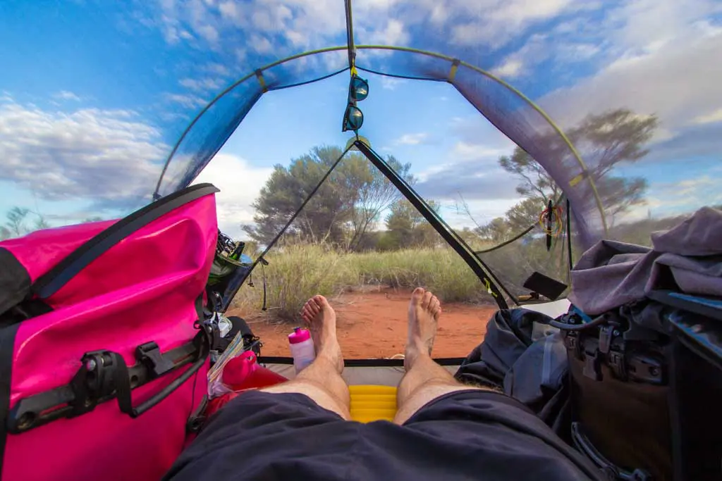 Person lying inside a tent with legs outstretched, surrounded by camping gear, including a pink bag. The tent is pitched in a bush camping spot with red soil, sparse vegetation, and a bright blue sky with scattered clouds.