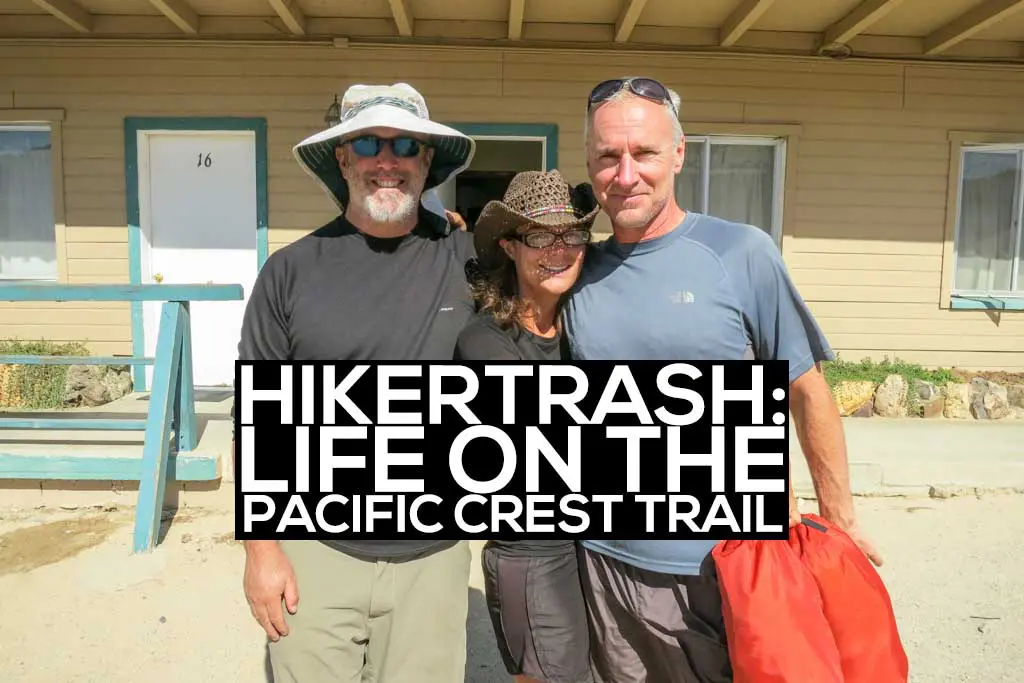 Three people stand closely, smiling, in front of a beige building. One wears a wide-brimmed hat, another a cowboy hat, and the third holds a red bag. A fitting ensemble for the Hikertrash life celebrated on the Pacific Crest Trail.