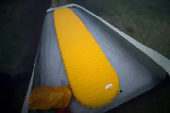 A yellow NeoAir XLite sleeping pad is placed inside a gray tent on a grassy surface. A yellow bag is positioned near the head of the pad. The tents interior appears dimly lit, suggesting an overcast day.