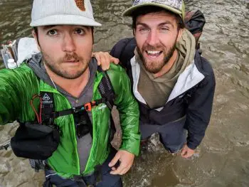 Two men stand knee-deep in water, posing for a selfie. One sports a green Montbell Tachyon Jacket and a white cap, the other a black and gray jacket with a patterned cap. Both have backpacks and are smiling against the riverbank backdrop.
