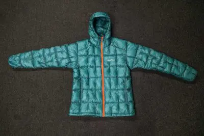 The Montbell Plasma 1000 Alpine Down Parka in teal, with its hood and striking orange zipper, lies flat on a dark surface. This puffer jacket showcases a quilted design and has its long sleeves outstretched.