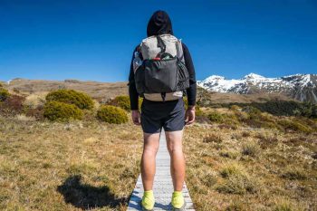 A person in a hoodie and shorts stands on a wooden path facing snowy mountains, carrying a Hyperlite Mountain Gear 3400 Southwest backpack. Bright green shoes contrast the grassy landscape, dotted with shrubs under a clear blue sky.