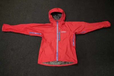 The Montbell Torrent Flier Jacket, a striking red with blue zippers, is laid flat on a dark carpet. It boasts a hood and long sleeves, complete with a logo on the left chest area, ensuring both style and functionality in any weather.