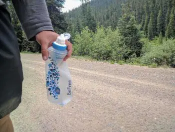 A person holds a Katadyn BeFree water bottle with blue designs on a dirt road, bordered by lush green trees and a dense forest. The path stretches endlessly ahead.