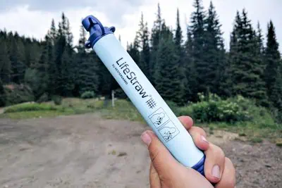 A person holds a LifeStraw with a forest backdrop, showcasing the portable blue water filter ideal for outdoor adventures. Its label features pictorial instructions, ensuring easy use in natures embrace.